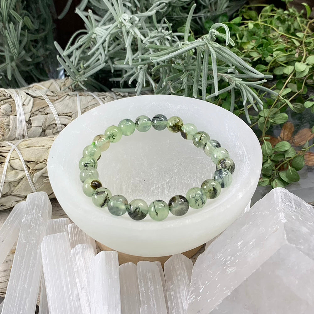 A prehnite mala bracelet of round pale-green beads with dark inclusions resting in a white selenite bowl