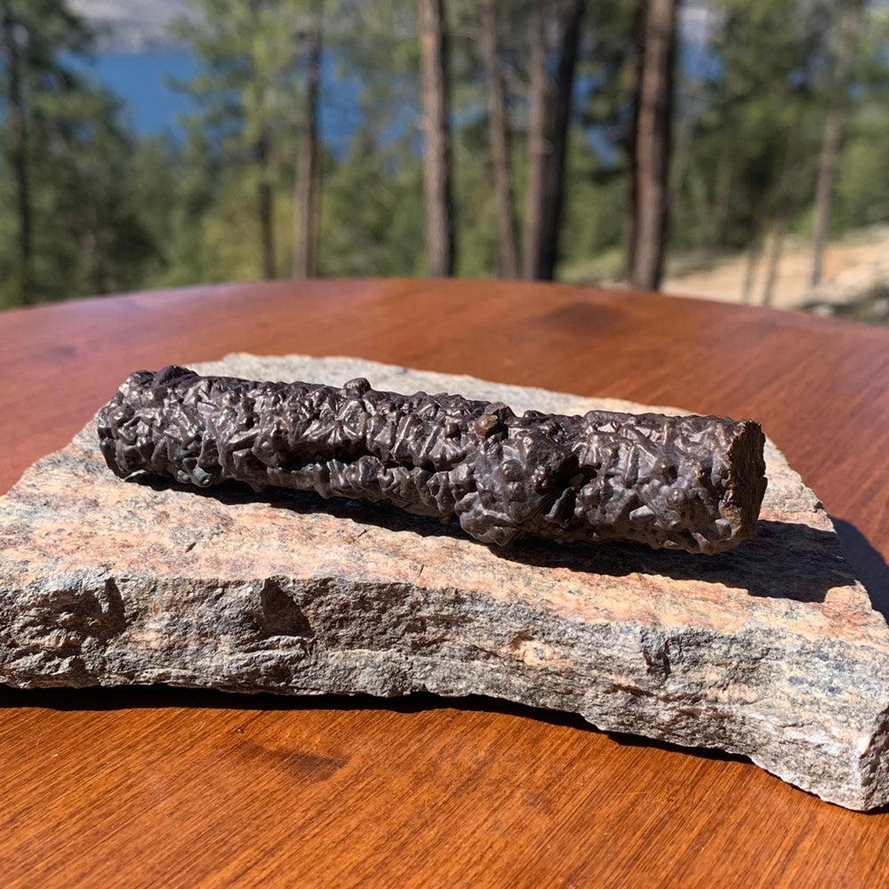 A rough, dark brown prophecy stone wand with a knobbly crystal-textured surface, lying on a slab of granite outdoors