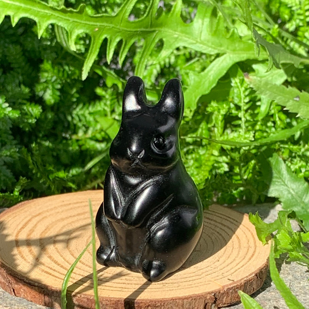 Glossy black agate rabbit totem sitting upright with tall ears on a wooden slice among green leaves in sunlight