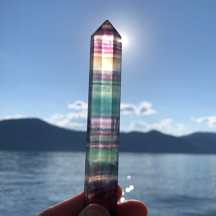 Rainbow fluorite point held up to the sunlight over a lake, its purple, green and gold bands glowing