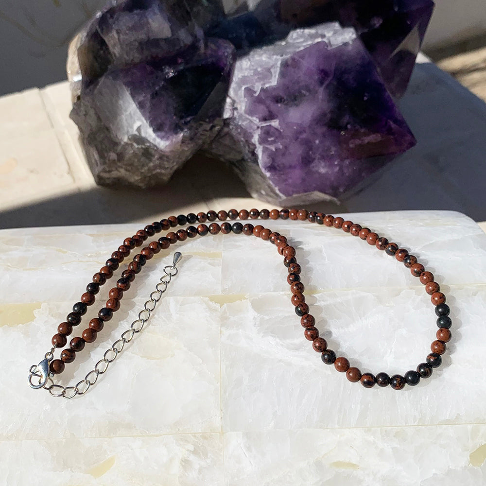 A red obsidian bead necklace in mahogany red and black, laid out with its silver clasp and extender chain on white stone