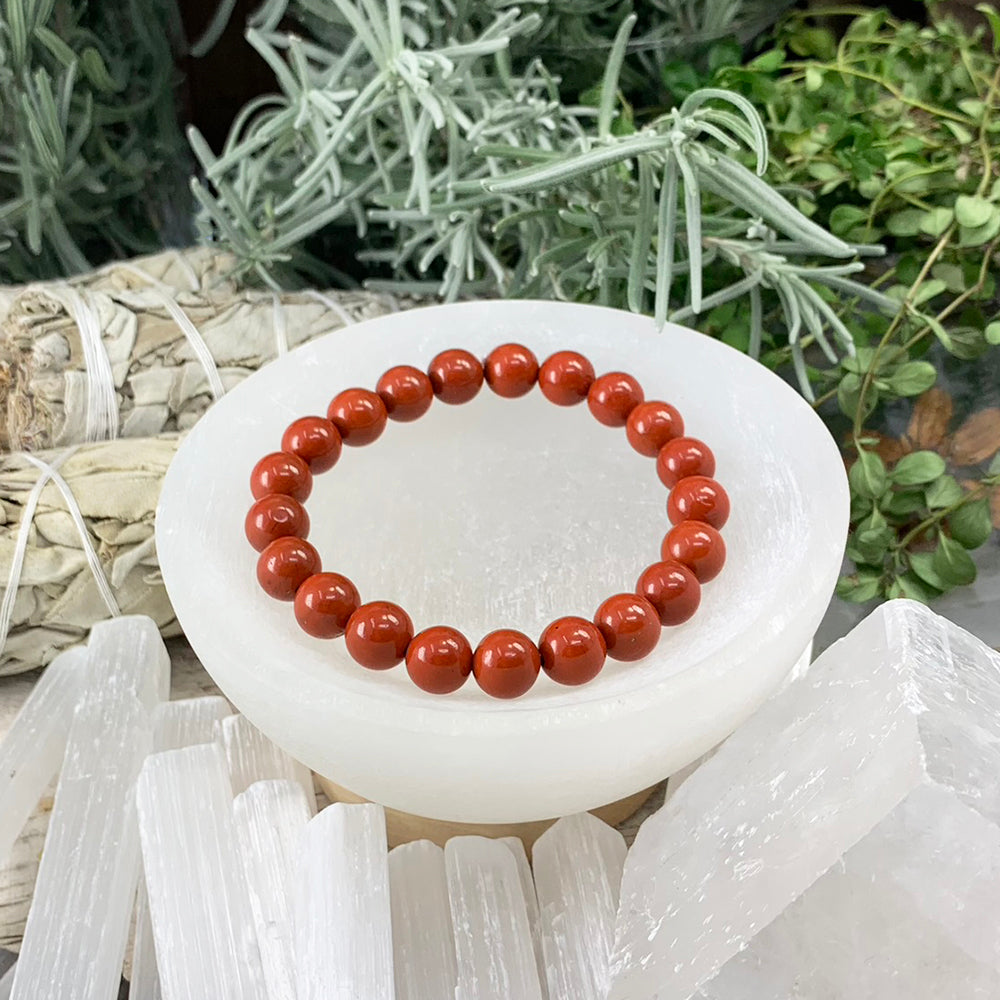 A single red jasper mala bracelet of polished round brick-red beads resting in a white selenite bowl