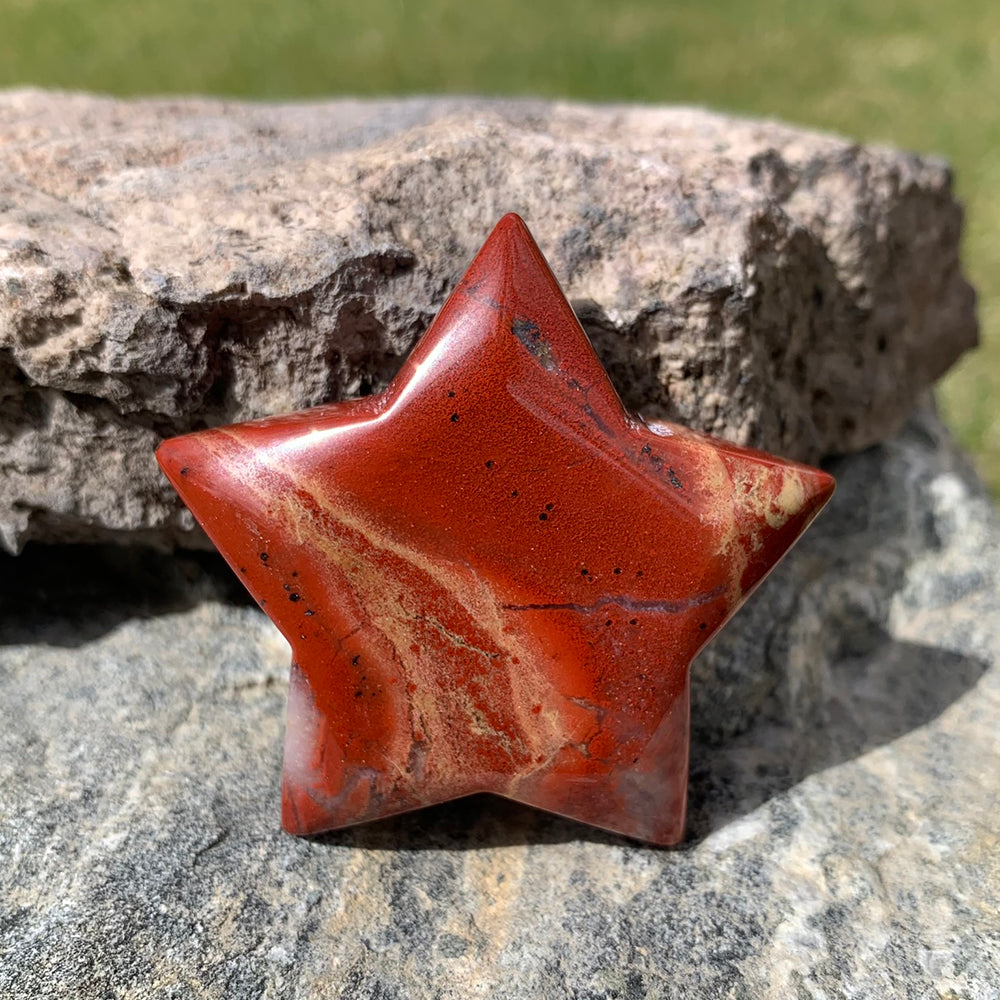 A polished five-pointed red jasper star in deep crimson with cream veining, resting on a rough rock in sunlight