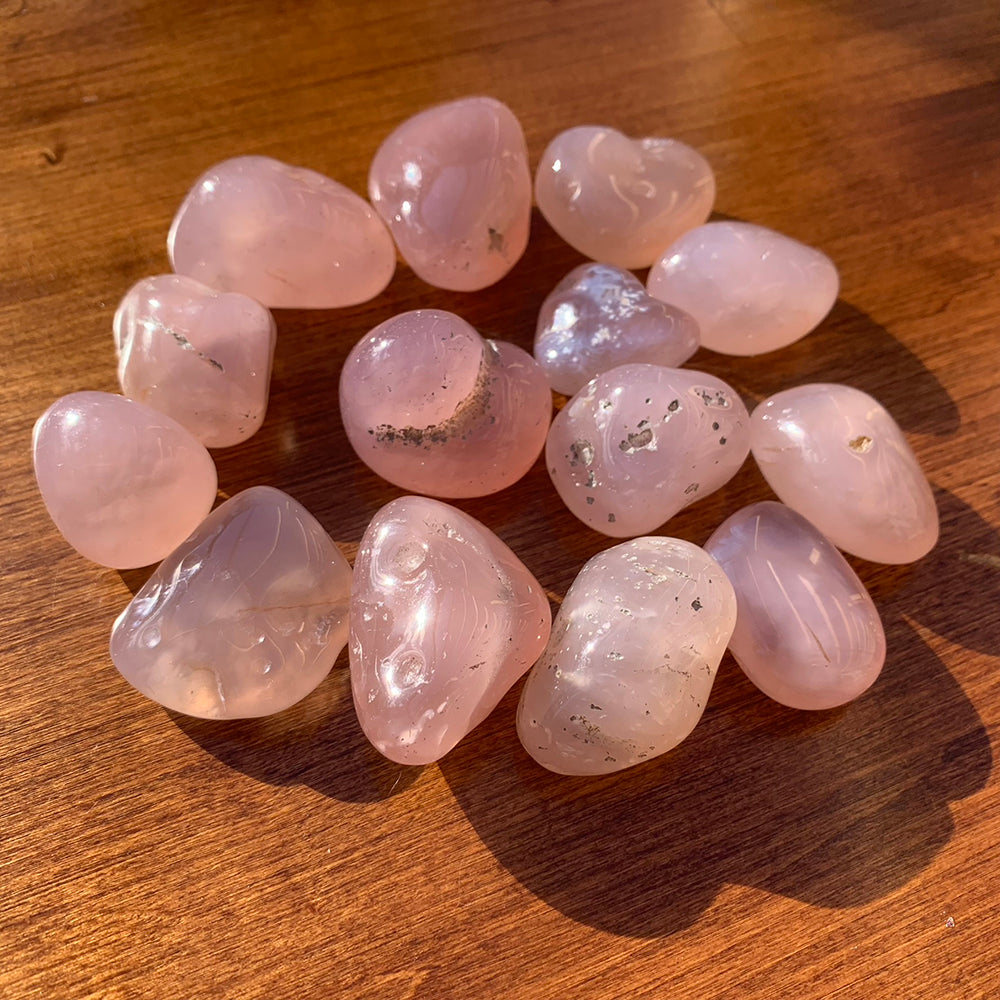 A circle of tumbled rose chalcedony stones in pale pink, several flecked with dark inclusions, on a wooden table in warm light