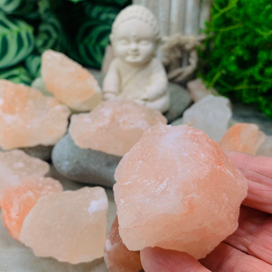 A hand holding a rough pink salt stone in front of more salt pieces, river stones and a small white Buddha figure