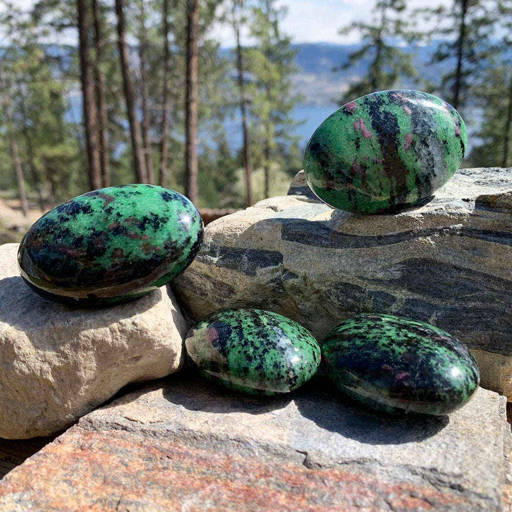 Four polished ruby in zoisite palmstones on sunlit rocks above a forest lake, each with its own scatter of ruby red through green
