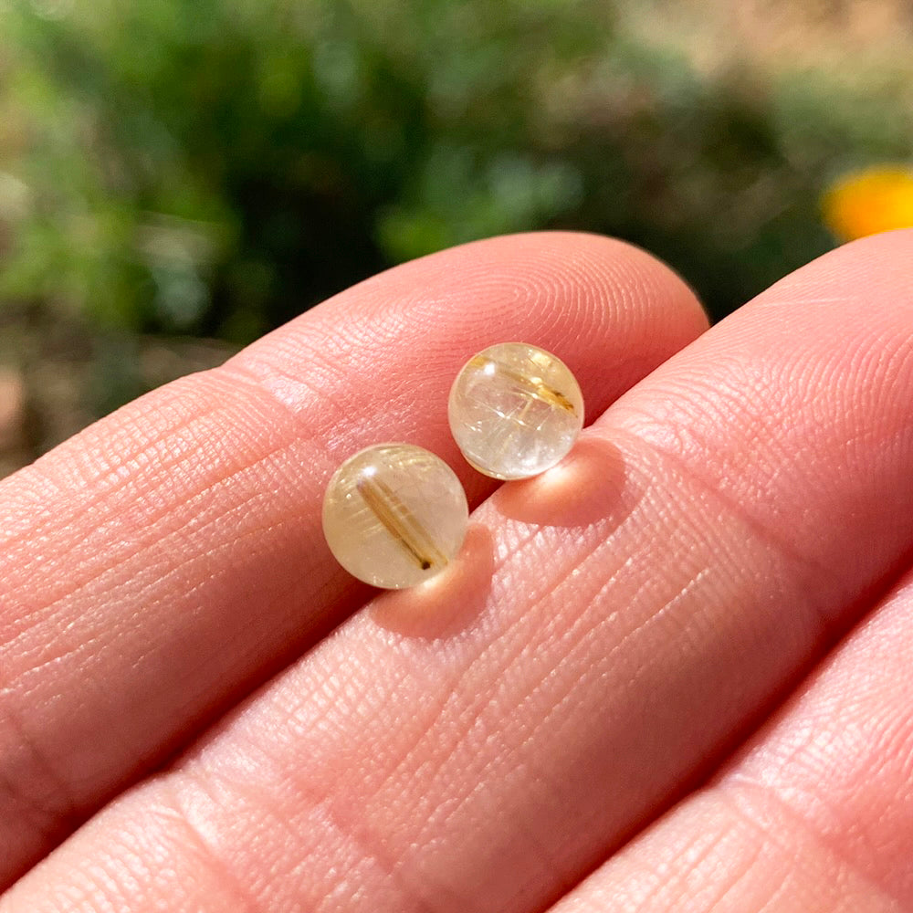 Two round polished rutilated quartz studs with fine golden needles inside, resting on fingertips in sunlight