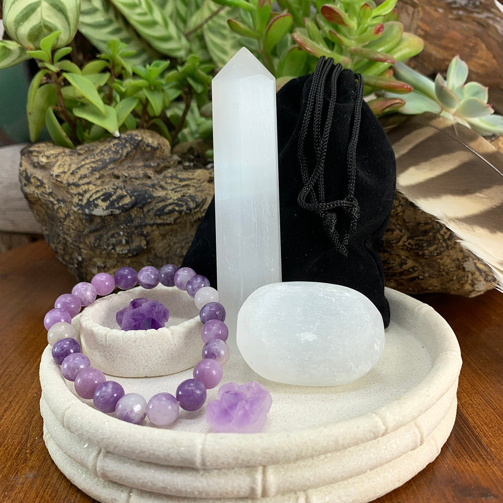 A selenite point, a selenite ellipse stone, a purple lepidolite bead bracelet and two small amethyst clusters on a white ceramic dish beside a black velvet pouch