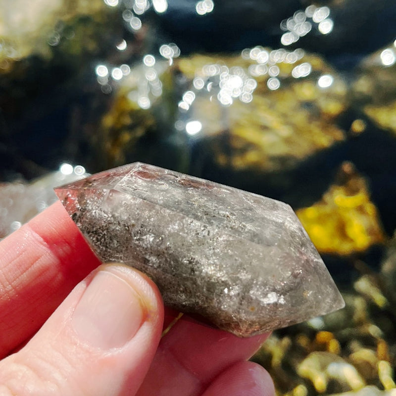 A double-terminated Shaman Quartz crystal held between thumb and fingers, showing dense gray inclusions
