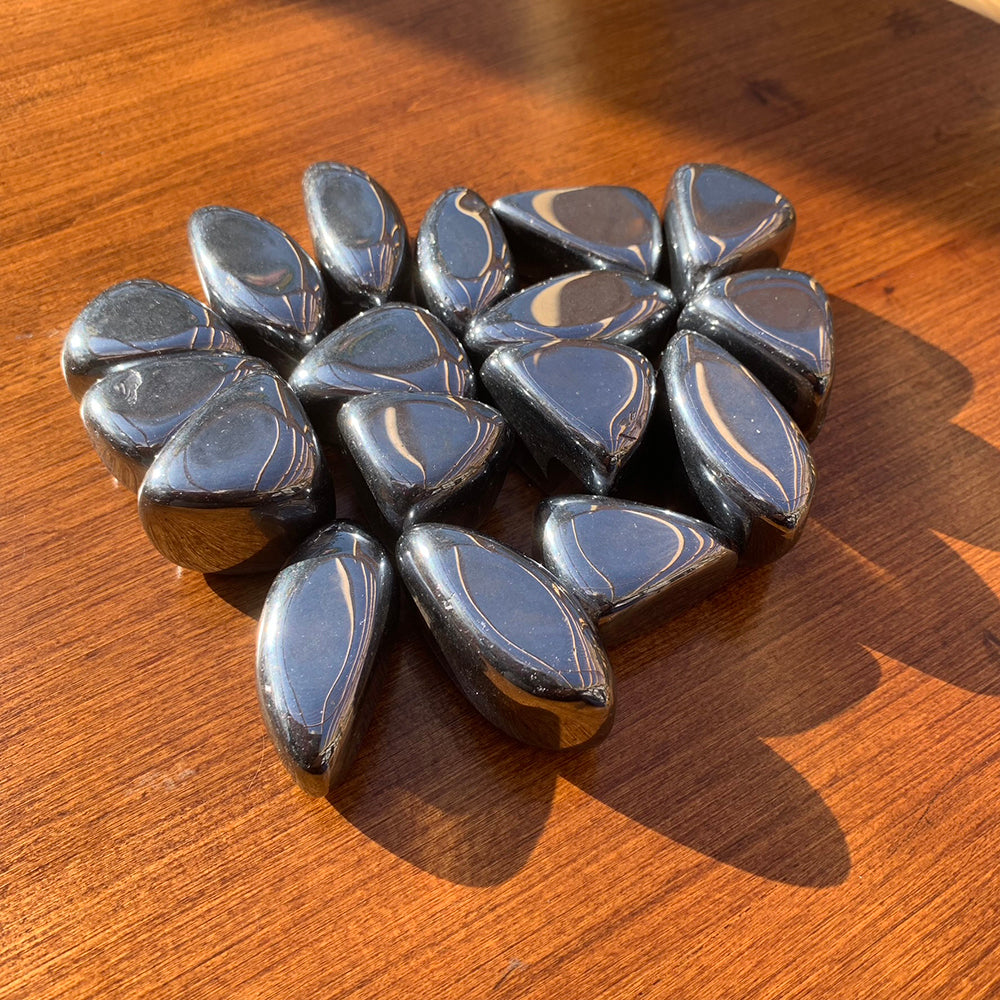 A cluster of sunlit hematite tumbled stones on a wooden table, each a slightly different teardrop or oval shape