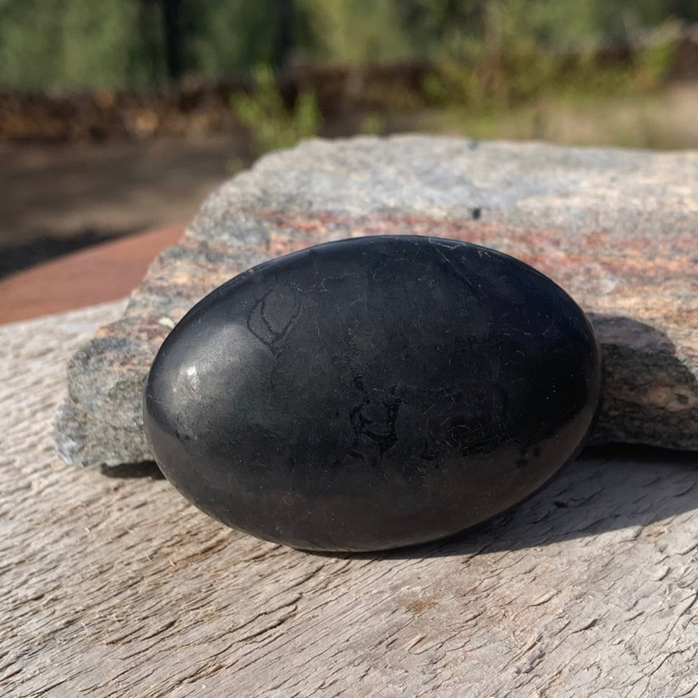 A polished black shungite palmstone resting on weathered wood and stone in the sun, its surface showing a soft metallic sheen