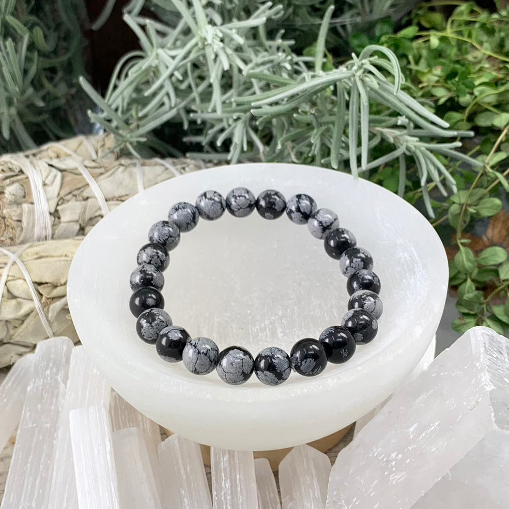 A snowflake obsidian bead bracelet, black beads flecked with grey-white snowflake patterns, resting in a selenite bowl