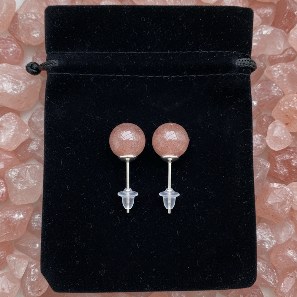 The pair of strawberry quartz studs with their posts and clear backs, laid on a black velvet drawstring pouch among rough strawberry quartz