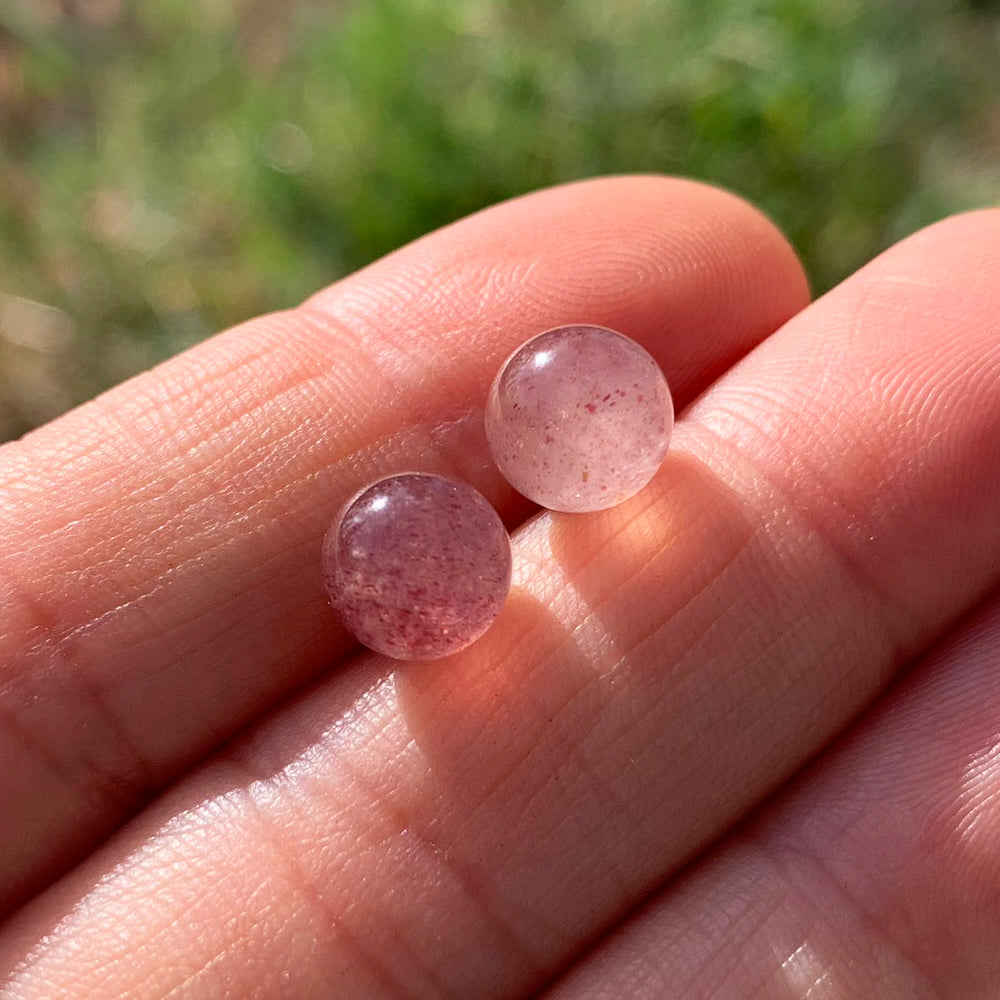Two round polished strawberry quartz studs with tiny red flecks, resting on fingertips in sunlight