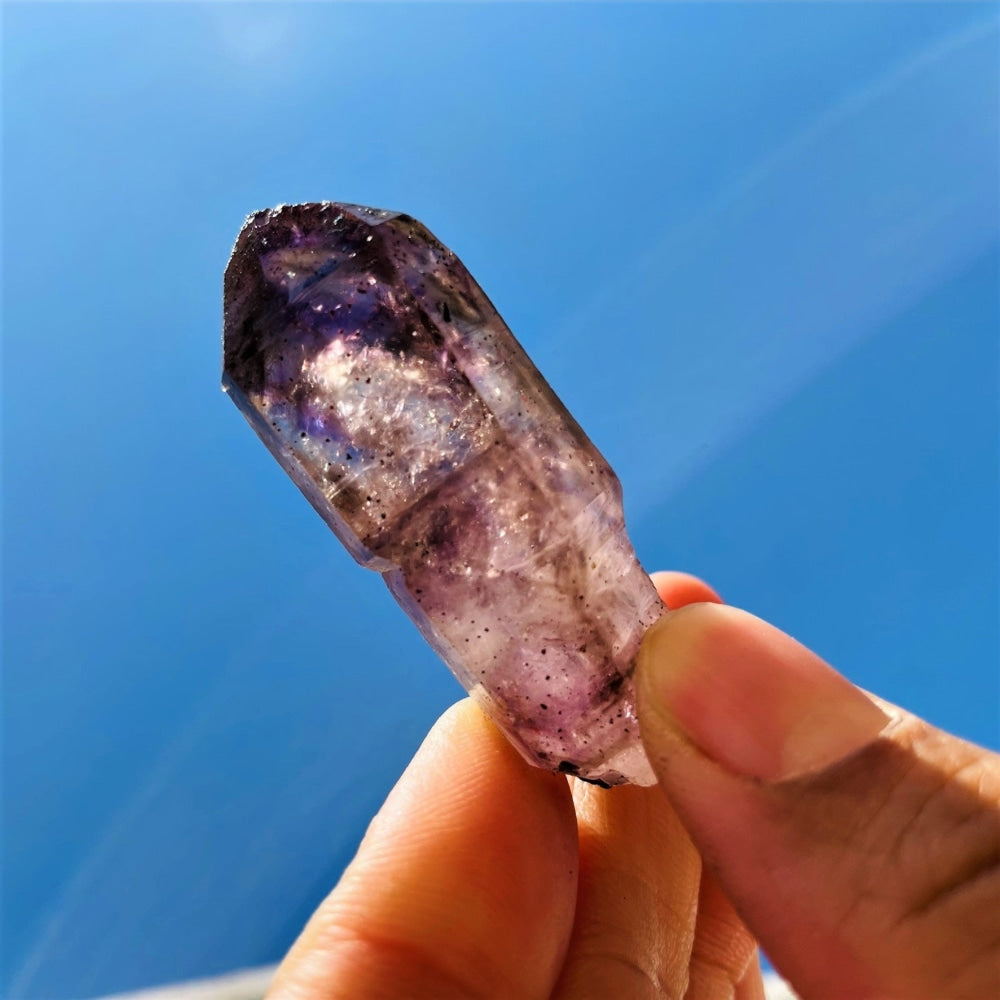 The small Super Seven point held up against a blue sky, smoky lilac quartz with a deep purple tip and fine dark inclusions